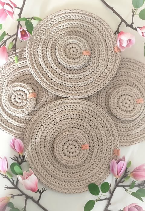 Four beige crocheted circular coasters arranged on white background with pink flowers and green stems
