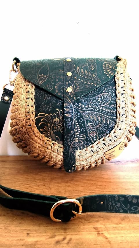 Dark teal patterned crossbody bag with tan and cream macramé trim on wooden surface with black strap