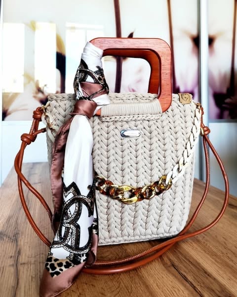 Beige woven bucket bag with wooden handles, cognac leather straps, gold chain belt, and decorative scarves draped over the side