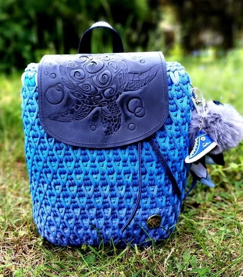 Blue knitted backpack with ornate dark blue leather flap on grass, small charm attached