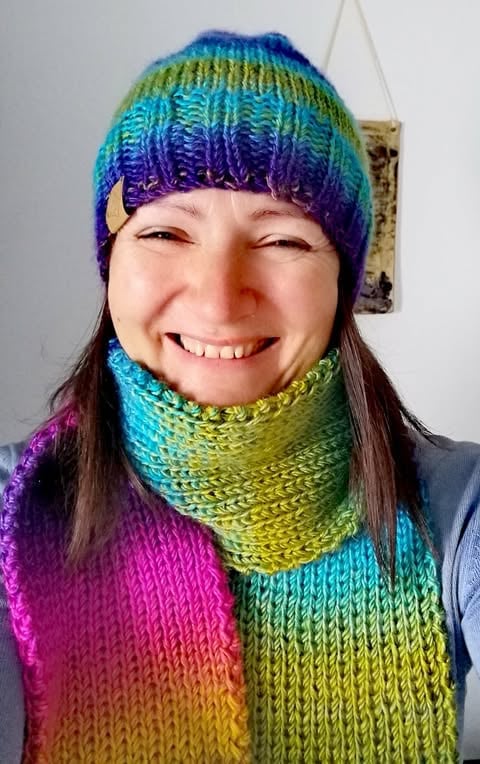 Smiling person wearing a colorful rainbow knitted beanie and matching scarf in shades of blue, purple, green, and yellow