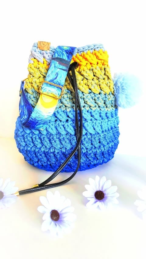 Colorful crocheted drawstring pouch in blue, yellow, and white with buckle closure and pompom charm, surrounded by white daisies